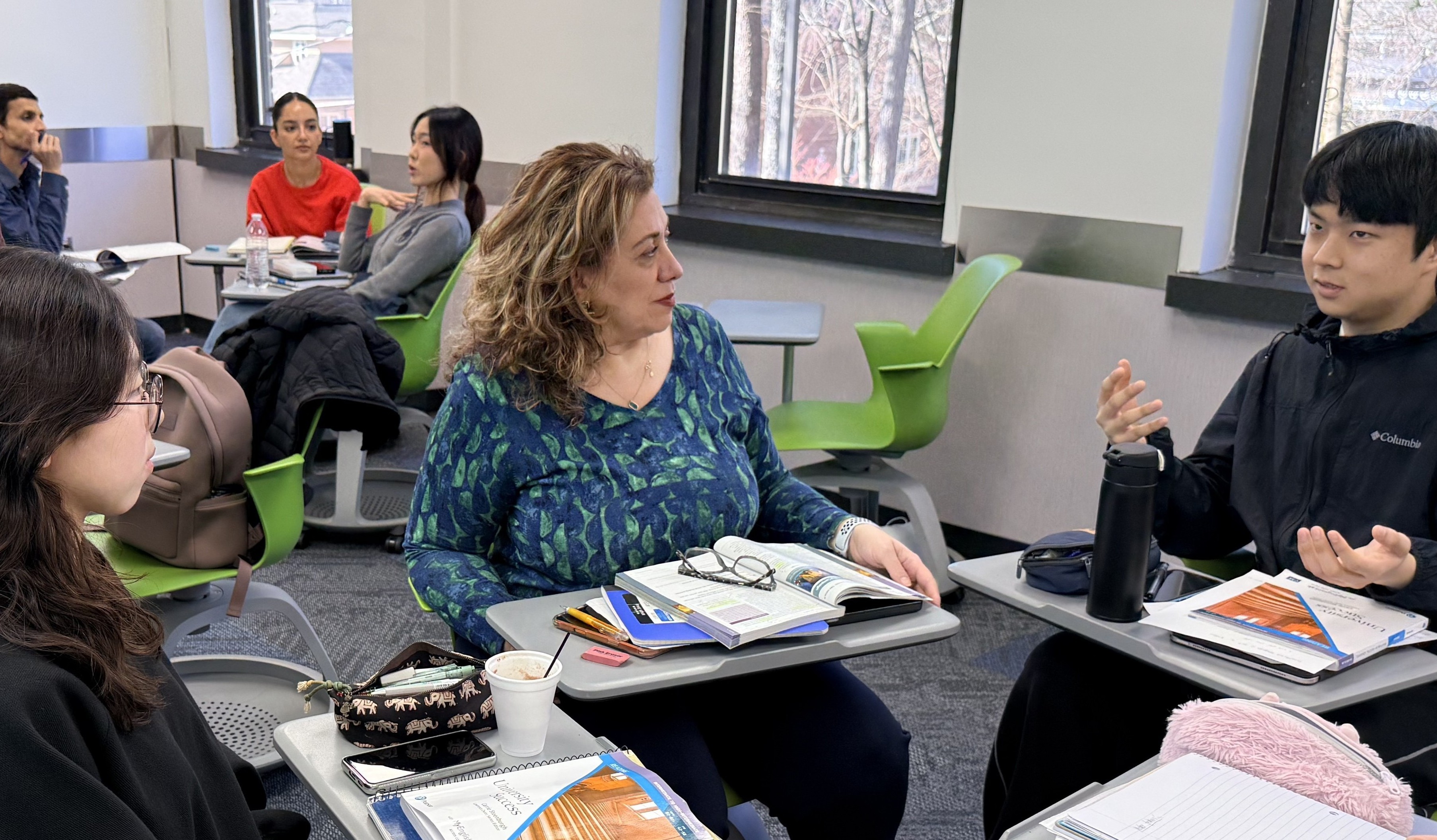 Students sit in small groups in a classroom, talking and working with notebooks and materials at their desks.